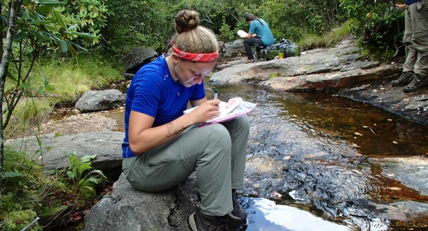 A person sits on a rock near a stream and journals 